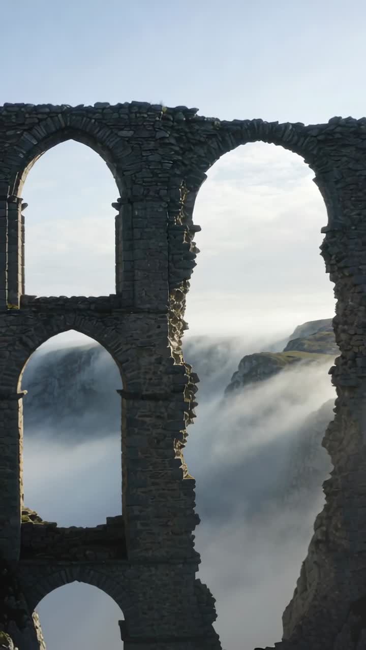 Vidéo verticale : Caméra reculant révélant trois ruines arquées à flanc de falaise, avec du brouillard dérivant.
