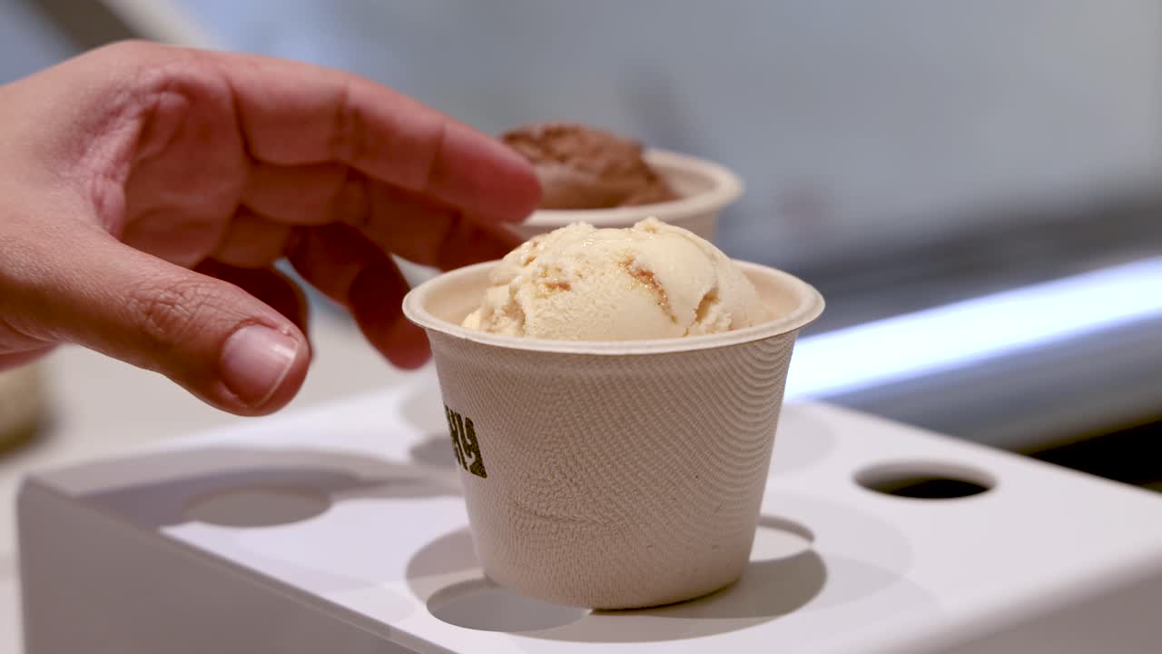 A person selects and lifts a paper cup of ice cream from a modern display stand under bright indoor lighting, with smooth camera movement and a clean background