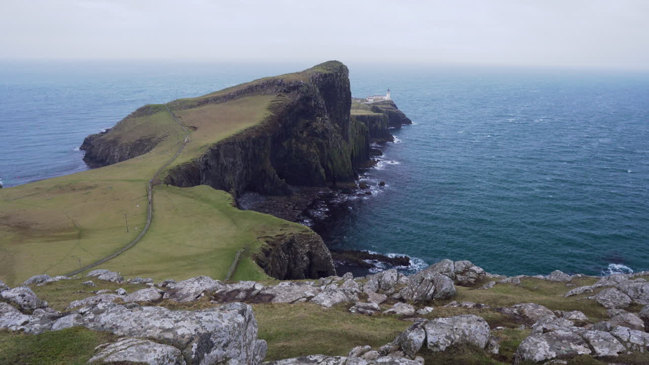 tiro inclinado hacia arriba del faro de neist point con acantilados rocosos en primer plano y el océano atlántico en el fondo en un día ventoso y nublado en escocia, isla de skye