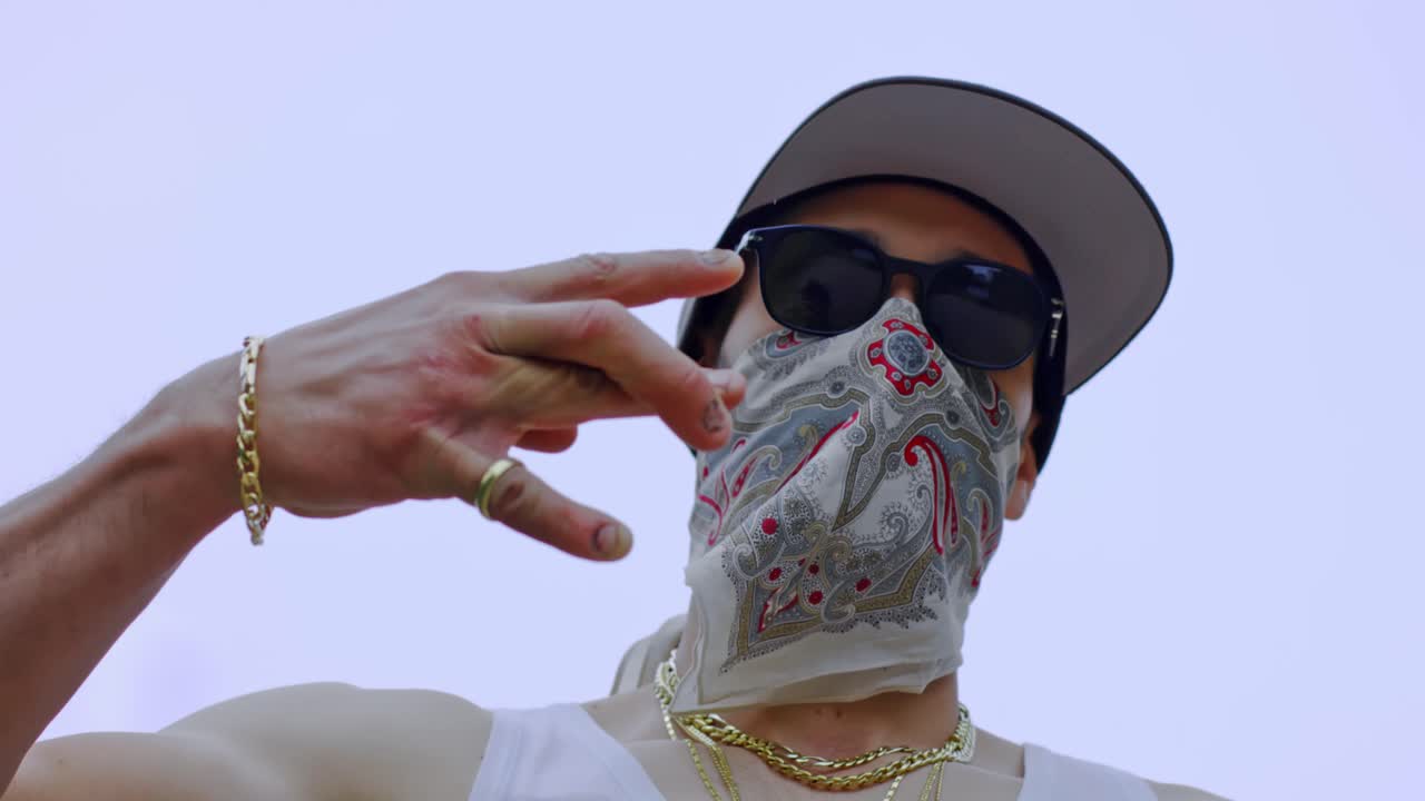 hombre adulto con collares de oro alrededor del cuello y bufanda sobre la cara hace w con la mano