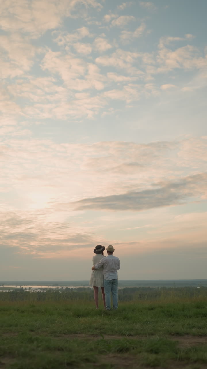 신혼 부부는 해가 지는 동안 조용한 호수 에 있는 잔디 언덕에 서서 하늘을 쳐다보고 있다. 색 셔츠, 모자, 청바지를 입은 남자는 여자의 허리를 잡고 있다. 그녀는 검은 모자와 색 드레스를 입고 있다.