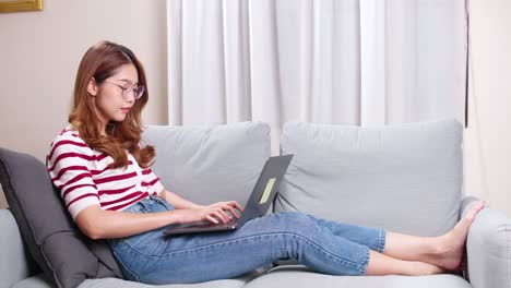 Young-Asian-woman-sitting-on-sofa-using-notebook-to-shop-online