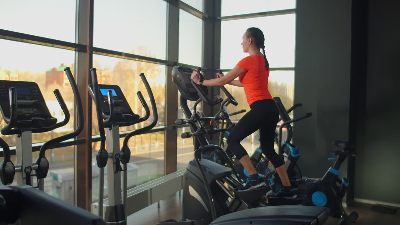 Cute young brunette doing thigh exercises on the simulator. Modern simulators near the panoramic window.