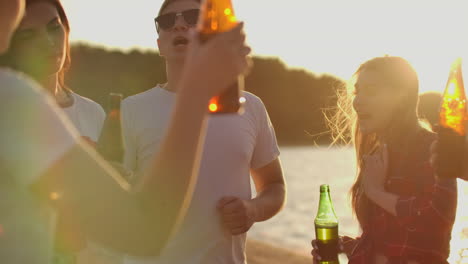 group of students celebrate the end of the semester with beer and pop music on the beach. they are dancing on the open air party at sunset in hot summer evening.