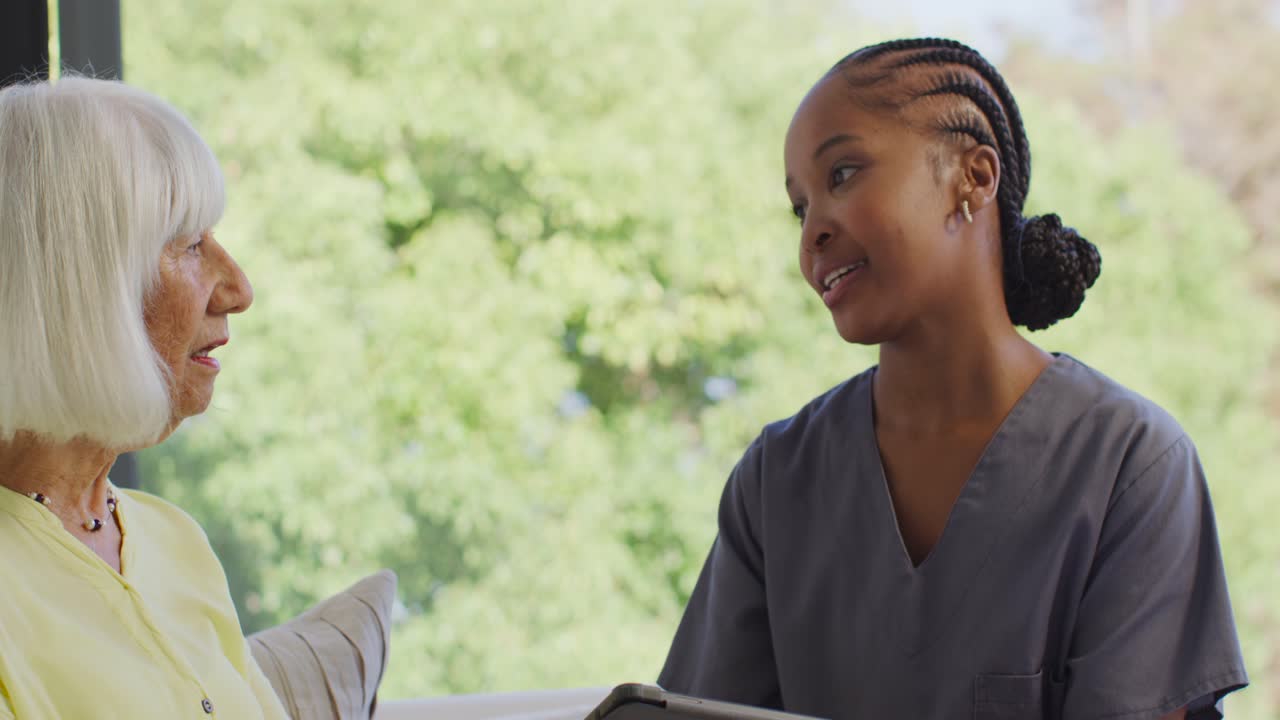 Female caregiver in scrubs holding tablet, showing screen and confirming info with senior on couch