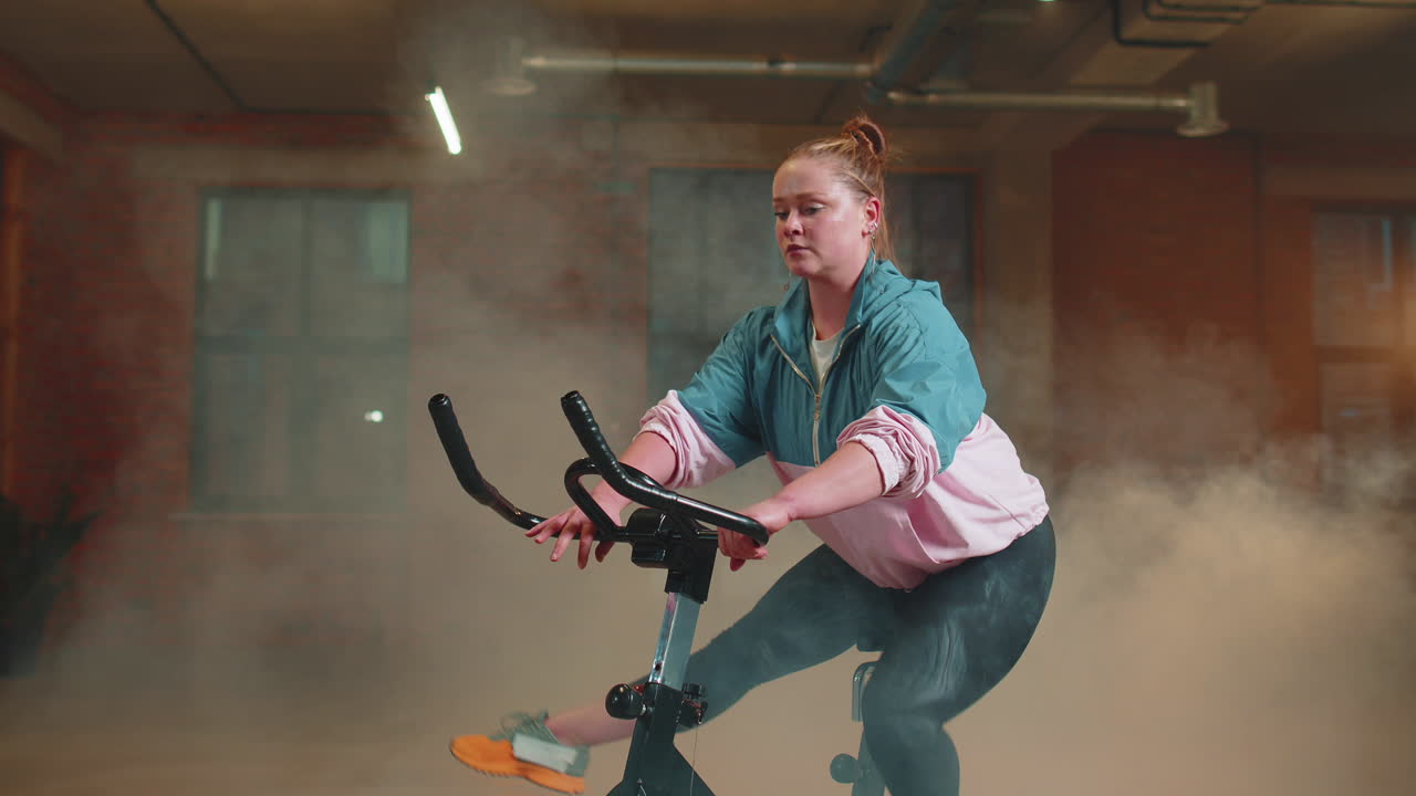 chica atlética realizando ejercicios de entrenamiento de equitación aeróbica en bicicleta estacionaria en un gimnasio con niebla