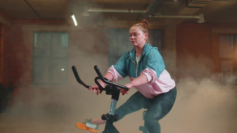 Athletic-girl-performing-aerobic-riding-training-exercises-on-cycling-stationary-bike-in-foggy-gym