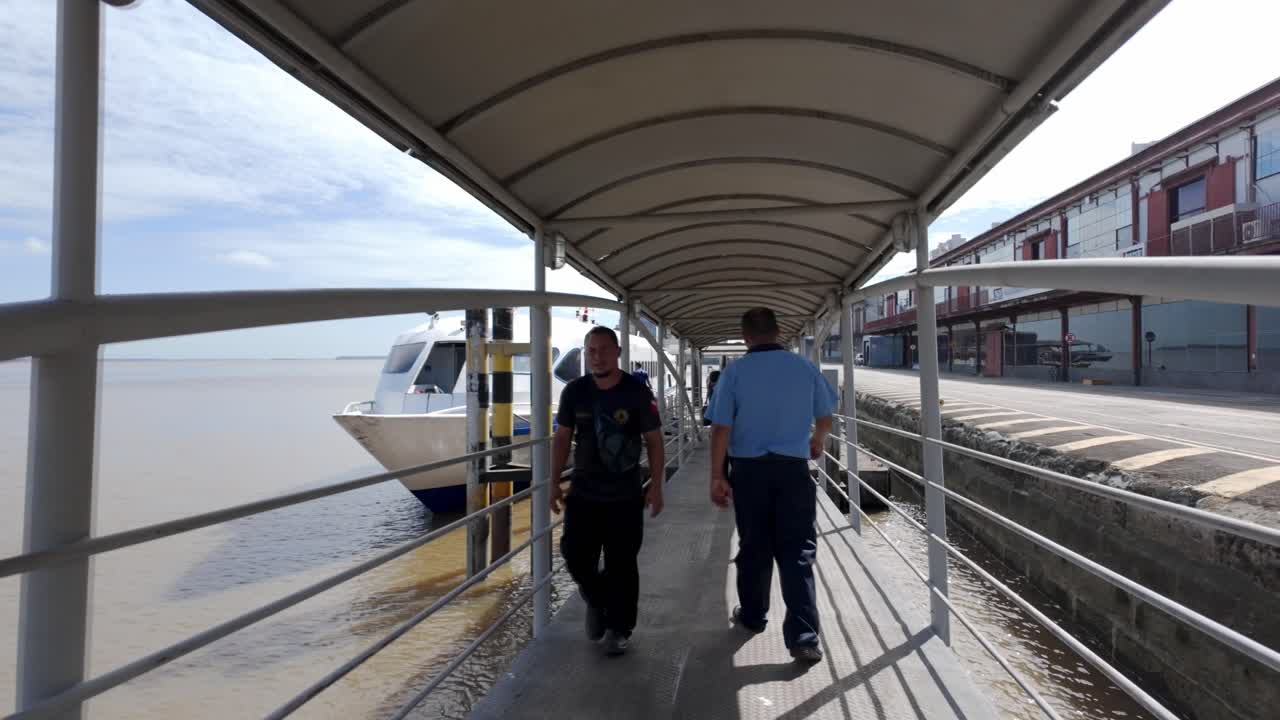 People walking on a covered pier towards a docked ferry boat at a port