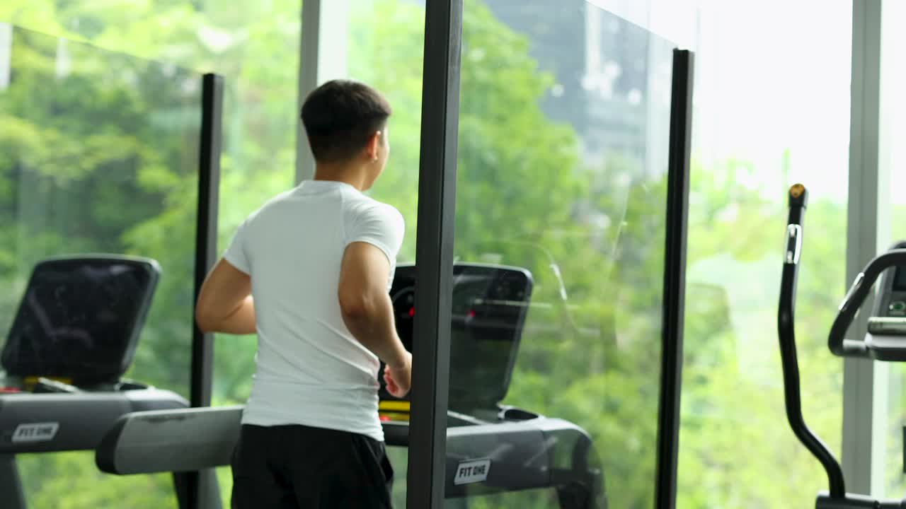 A man exercises on a treadmill in a modern gym with large windows overlooking greenery. Bright, natural lighting enhances the scene