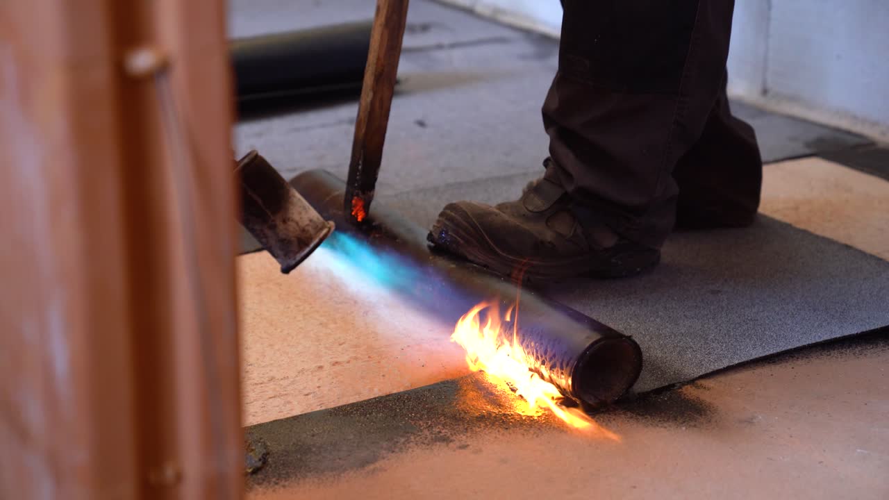 Worker uses gas torch to heat and seal bitumen sheet on concrete floor during construction
