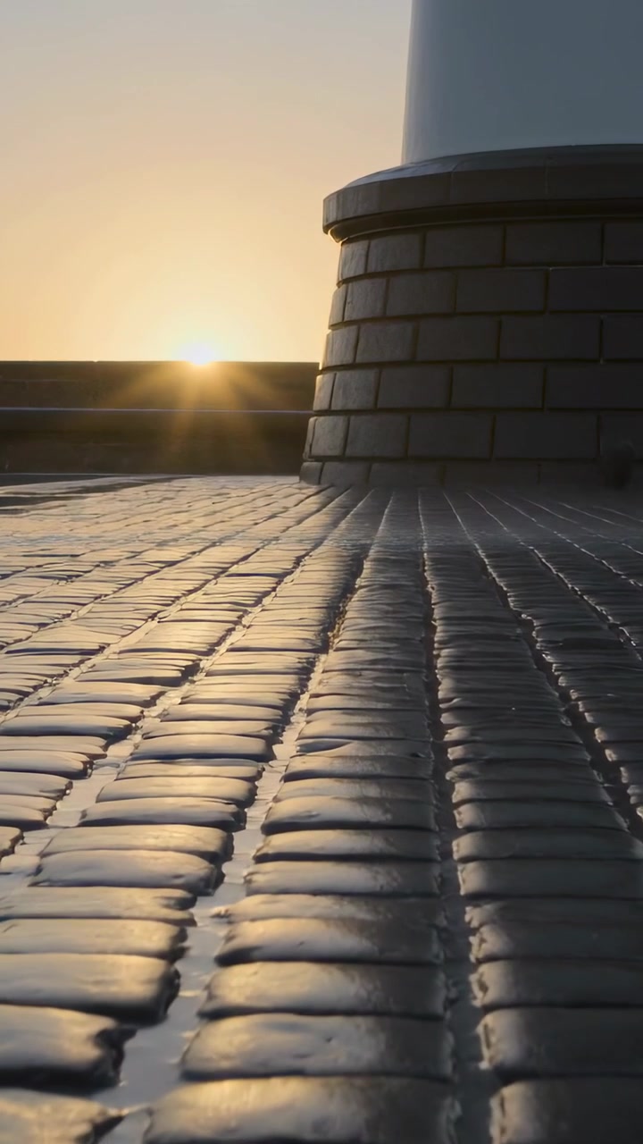 Vertical video: Sun rising, wet cobbles reflecting glints to horizon, mist drifting by shore tower