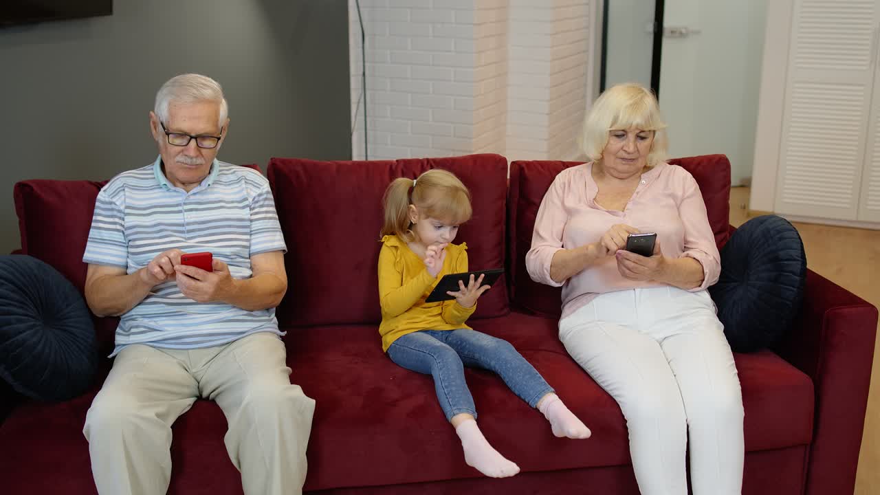 Bedsteforældre og barnebarn sidder i sofaen derhjemme og bruger mobiltelefoner og digital tablet.