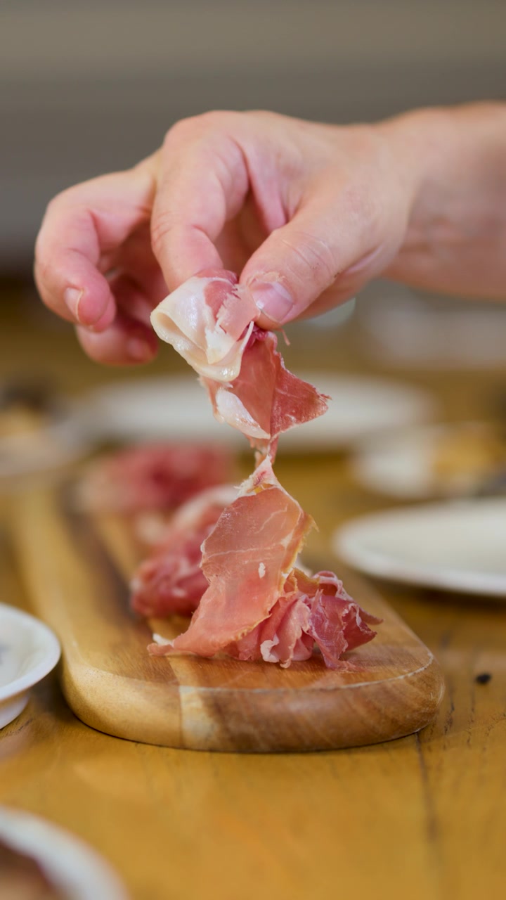 Una mano prende sottili fette di prosciutto crudo da un tagliere di legno durante la cena