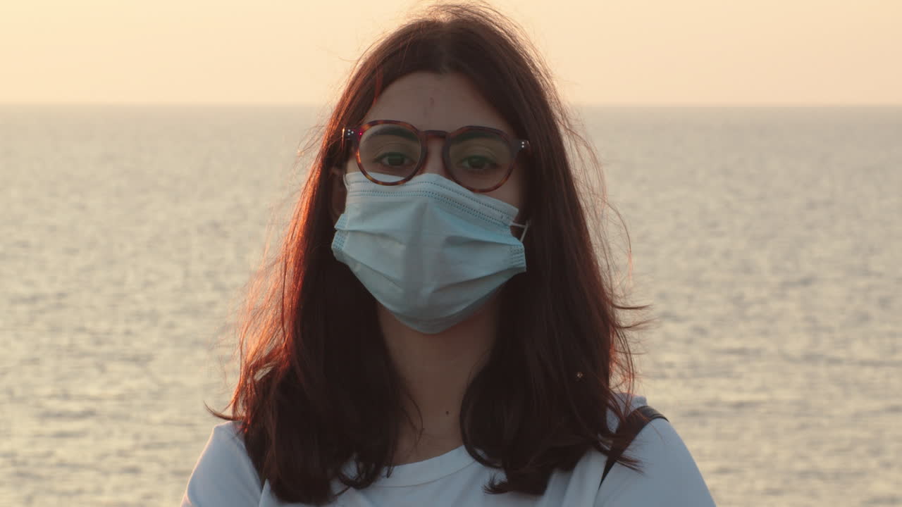 Vista frontal de una joven europea con mascarilla mirando a cámara con el mar de fondo, desesperanzada sobre el futuro de la pandemia de COVID-19, estática, primer plano