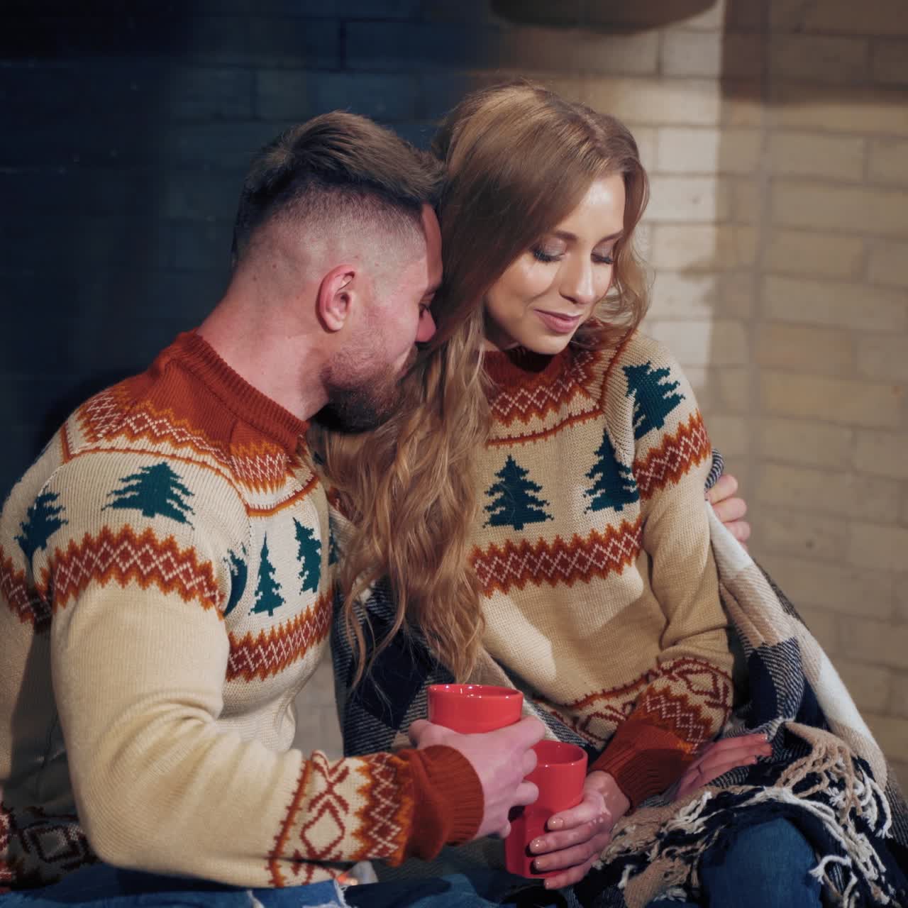 Beautiful loving couple resting near fireplace. Young woman and a man with cups of drink at Christmas. Guy hugging his girlfriend enjoying romantic weekend.