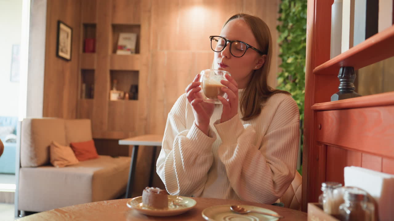 young woman in white sweater drinks latte from clear glass cup while seated at cafe table with chocolate dessert, warmly lit background with cozy furniture and decorative shelf