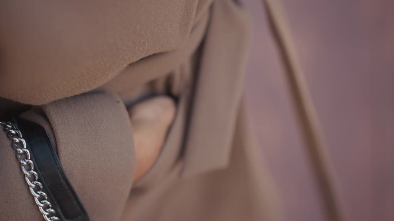 Primo piano di una donna che indossa abiti in tessuto marrone con una texture morbida e una tracolla a catena d'argento, concentrandosi sui dettagli del materiale. Il soggetto rimane immobile per tutto il tempo, enfatizzando la trama del tessuto.
