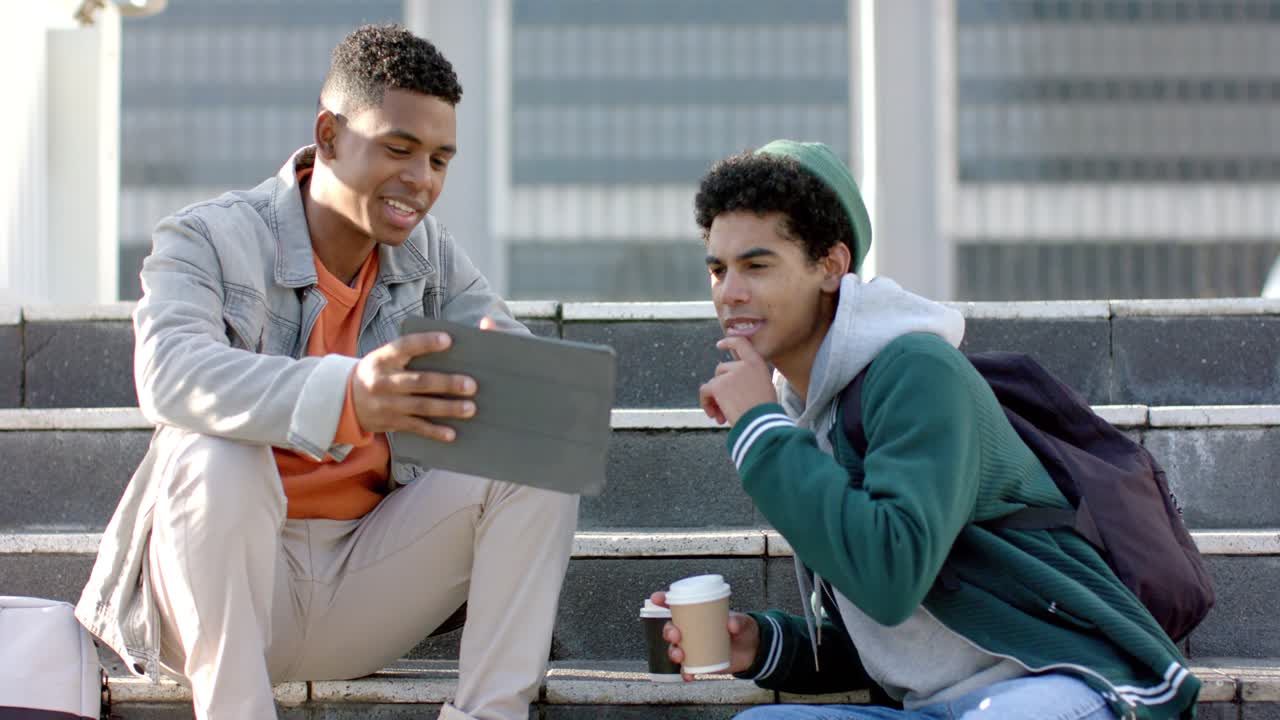 Amigos diversos do sexo masculino compartilhando um tablet enquanto seguram um café, revisando informações nos degraus de uma praça urbana