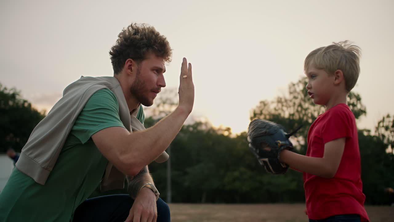 padre con cabello rizado y rastro en una camiseta verde ayuda a su hijo pequeño rubio en una camisa roja ponerse un guante de béisbol y alto-fives él en el parque