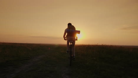 a biker with a backpack rides uphill along a rural road in the rays of the setting sun slow motion v