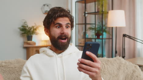 surprised indian bearded man using cellphone winning lottery sitting on sofa couch at home apartment