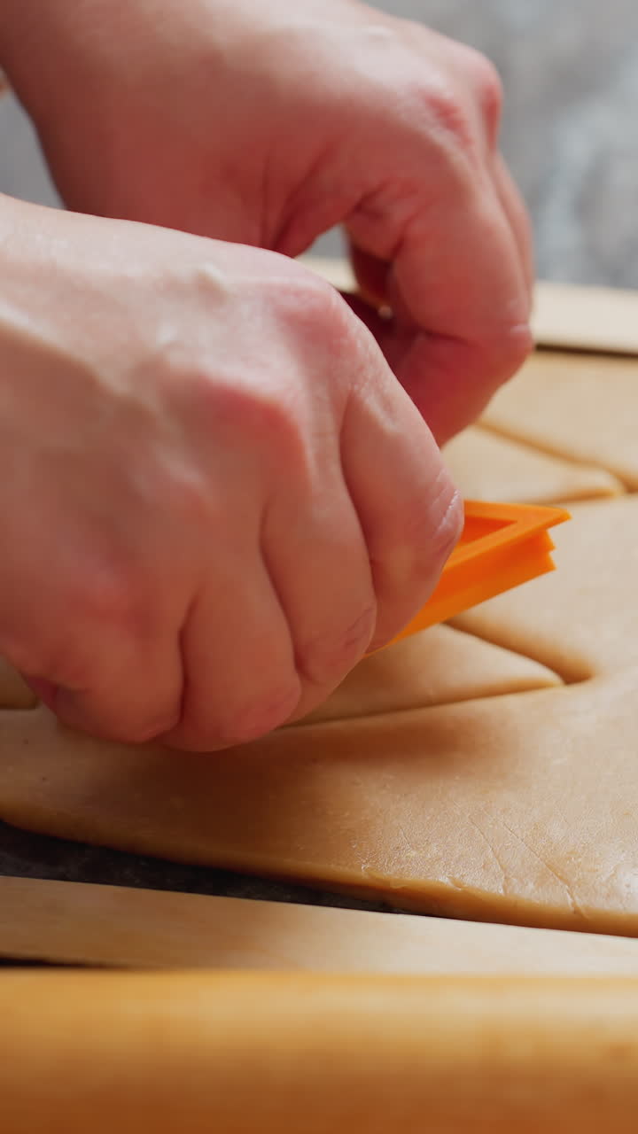 primer plano de manos de mujeres presionando un cortador de horneado naranja en masa marrón para crear formas precisas, rodillo de madera, herramientas de hornear y encimera texturizada en iluminación suave