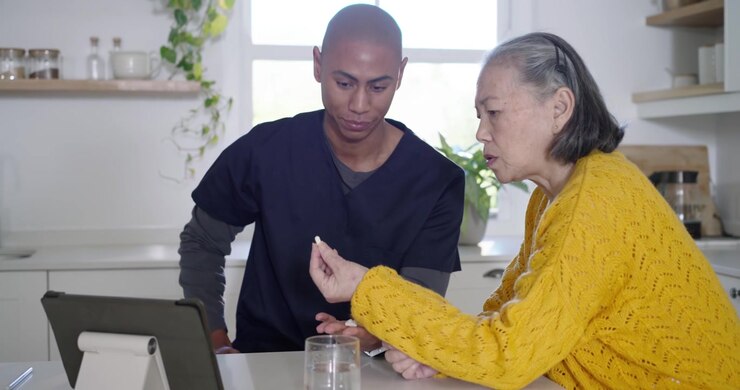 Senior Chinese woman in yellow pointing to tablet, African American aide checking meds at kitchen