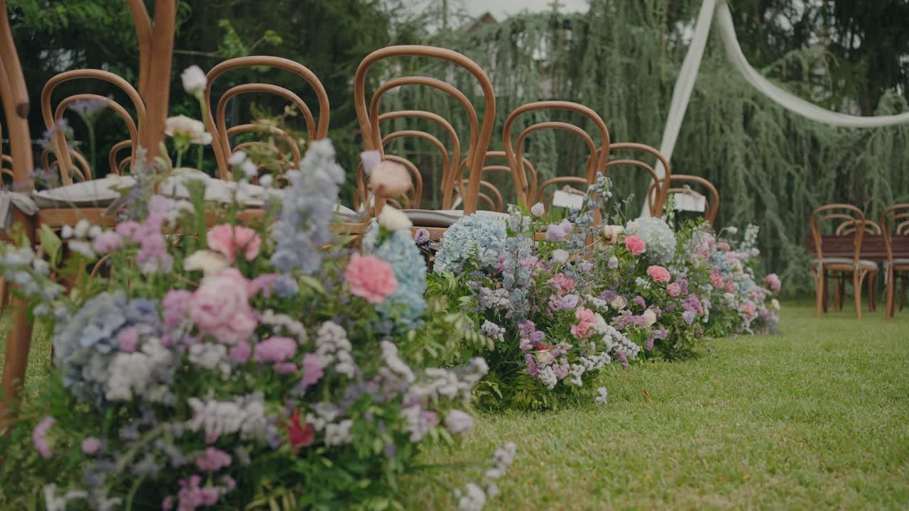 파스텔 핑크, 파란색, 라벤더 색상의 꽃들이 나무 의자가 배경으로 있는 야외 결혼식 통로를 장식하고 있어요.