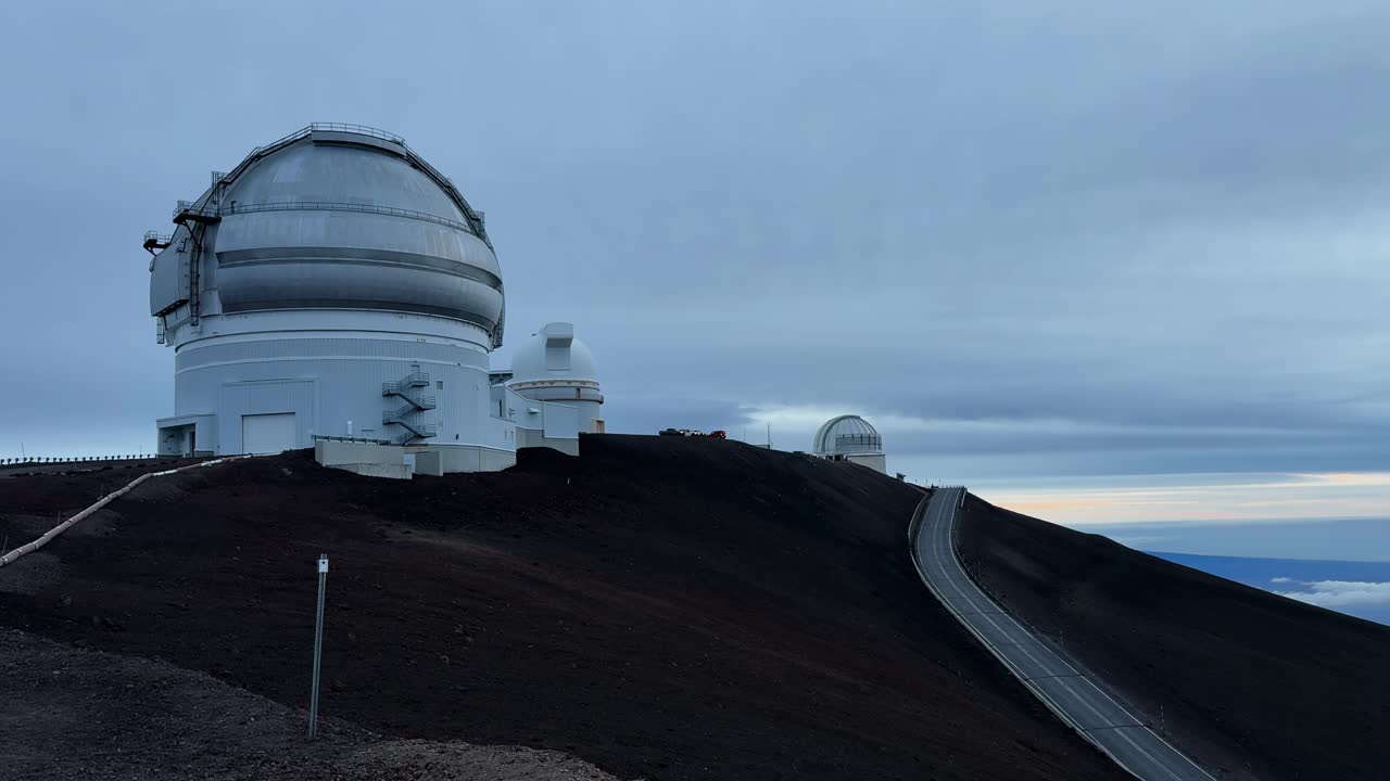 Static shot shows astronomical observatories on Mauna Kea with access road and dark volcanic soil