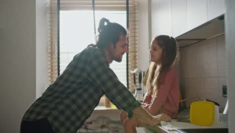 close-up shot of a brunette man in a green checkered shirt leaning on the table and talking with his little brunette daughter in a pink dress in a modern white kitchen