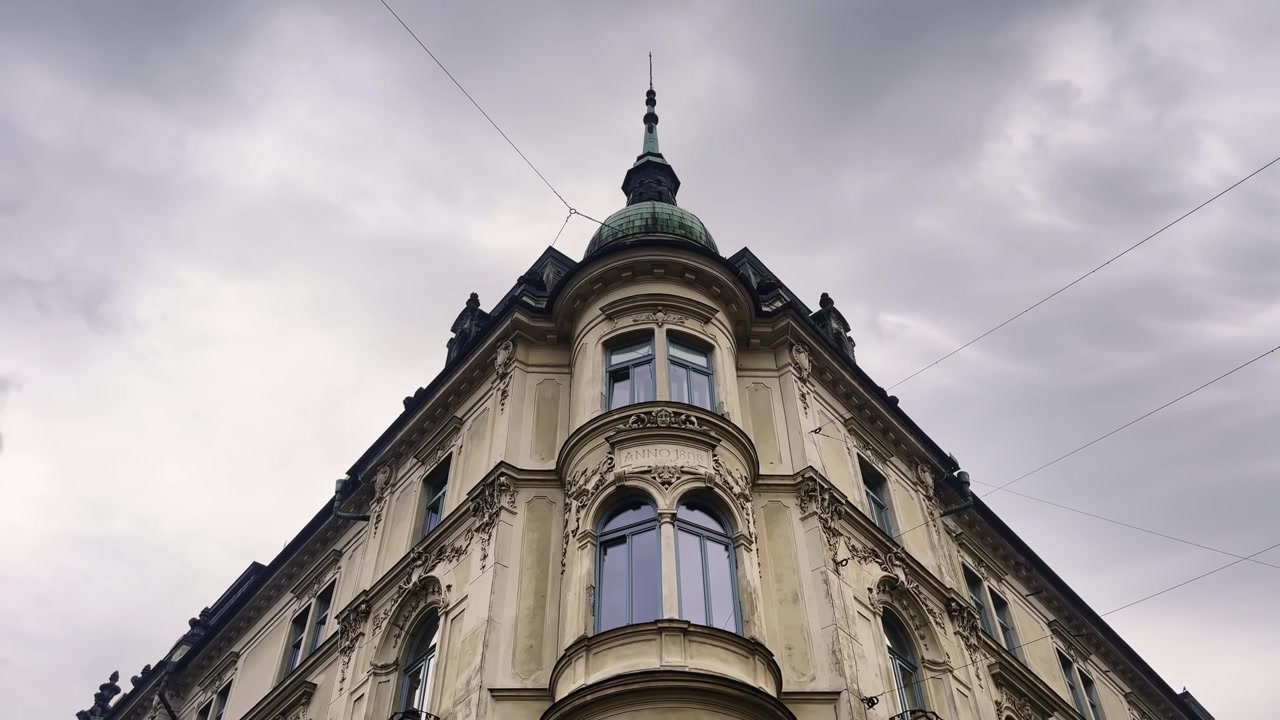 Vista de baixo ângulo de um edifício de esquina histórico ornamentado em Liubliana, apresentando uma sacada arredondada, janelas em arco, fachada esculpida e uma torre com cúpula de cobre sob nuvens dramáticas