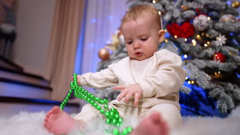 adorable one-year-old baby in white clothes sits playing with beads string. low angle view on the child focused on christmas decorations.