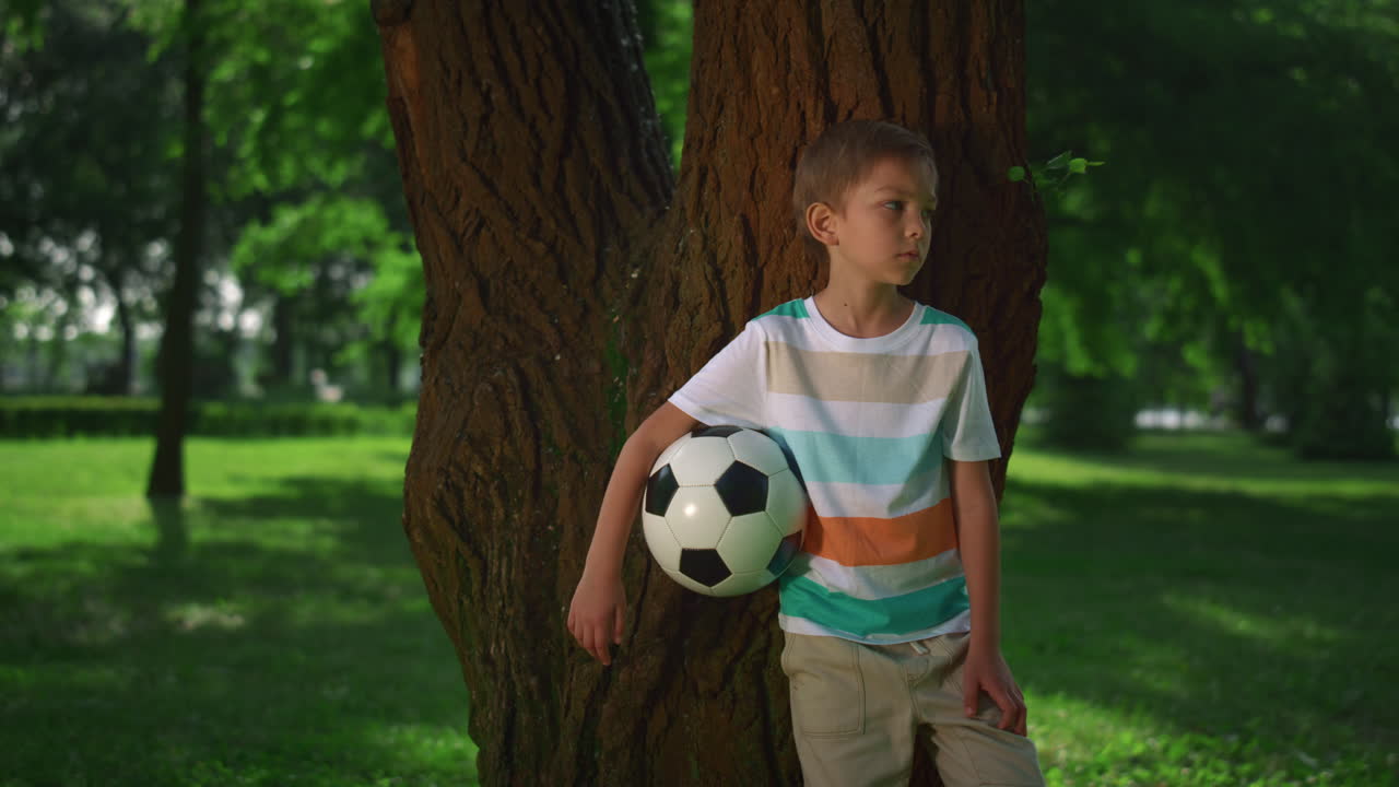 Menino pequeno segurando bola de futebol encostado na árvore. Jovem atleta posando com a bola em close-up.