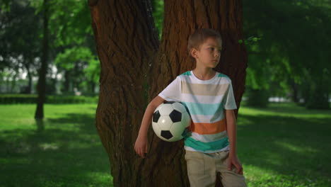 little boy hold soccerball lean on tree. young athlete posing with ball closeup.