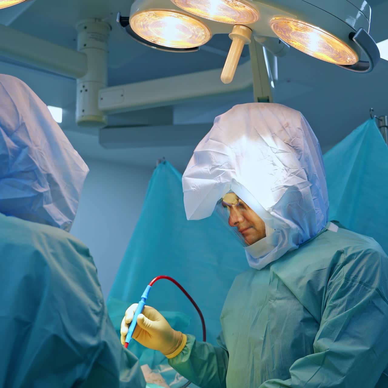 Professional team of surgeons in protective suits working under bright lamps in surgery room. Doctors use modern device at operation