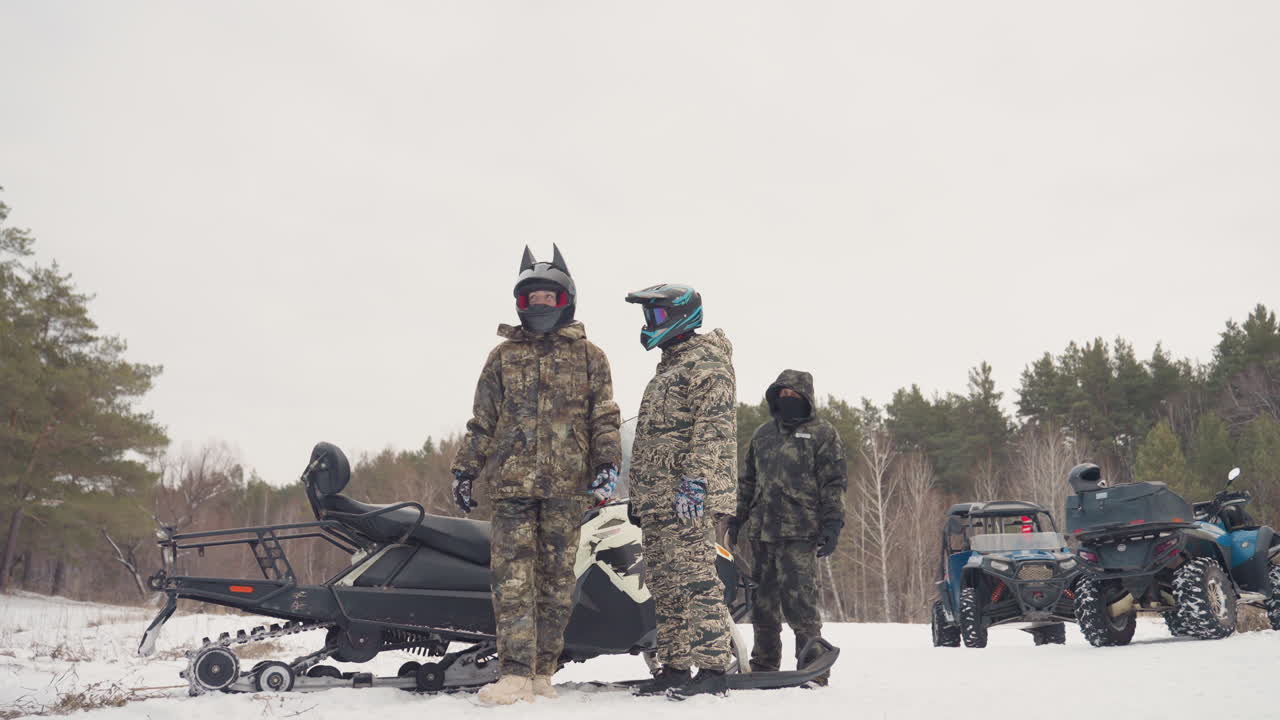 Il leader parte per esplorare il sentiero innevato oltre una motoslitta parcheggiata mentre i compagni osservano il suo passo solitario; l'orizzonte delimitato da pini, il cielo coperto e la quiete contemplativa definiscono il tono per l'esplorazione e il percorso.
