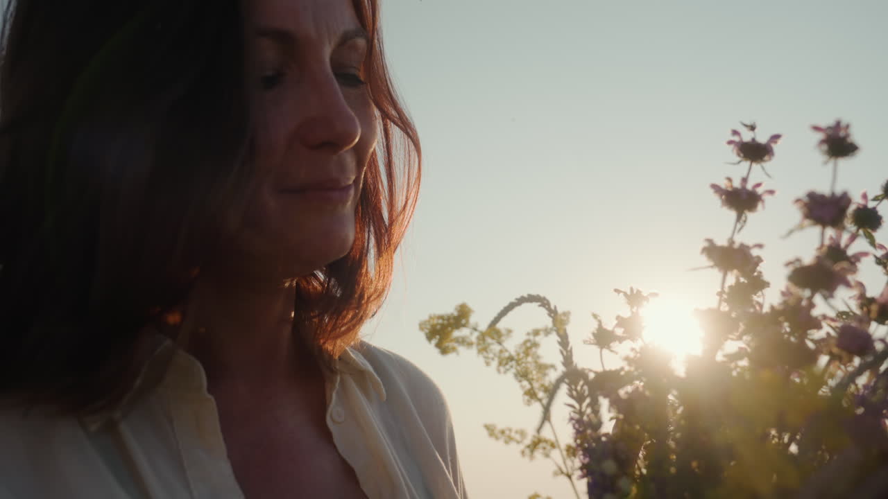 close up of woman with white hat and shirt holding wildflower bouquet in hand leaning forward to smell stalk of yellow bloom with grimace as sunlight backlights scene in grass field