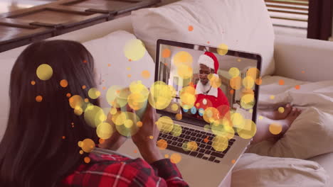 animación de puntos de luz sobre mujer biracial en llamada de video portátil con papá noel en navidad