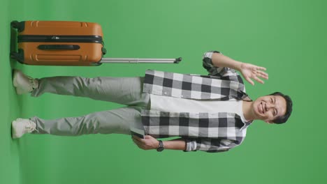 full body of asian male traveler with luggage and passport smiling, waving hand, and saying bye while standing in the green screen background studio