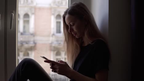 Mujer-Joven-Sentada-En-El-Alféizar-De-La-Ventana-Y-Usando-Un-Teléfono-Inteligente