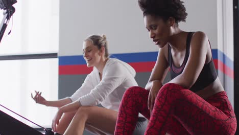 video of two happy diverse women on rowing machines recovering after working out at a gym