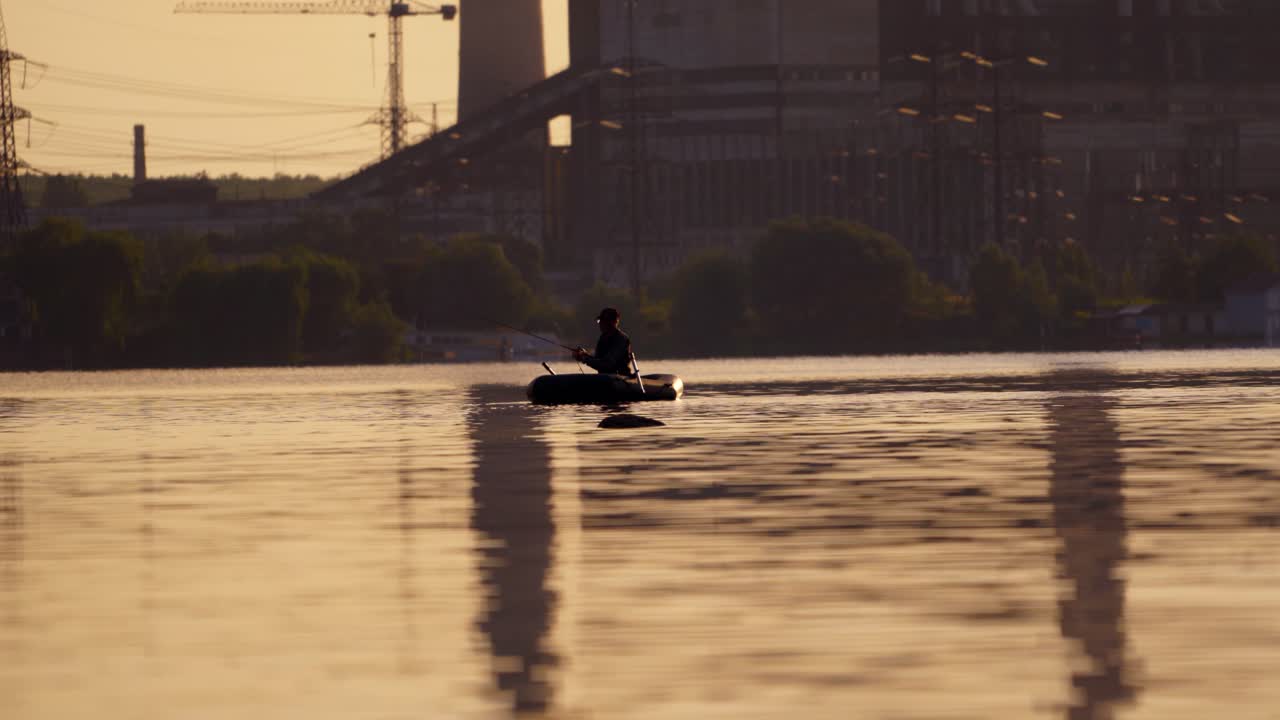 Fisherman in a boat with fishing rods in the evening. Man in hat floating in a boat and fishing on the background of a factory in the city.
