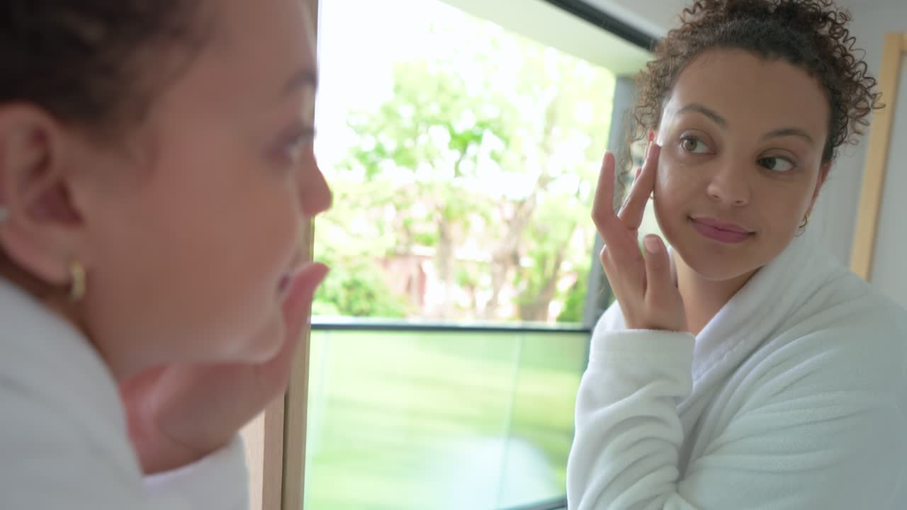 Donna afroamericana in accappatoio bianco che apre un barattolo e applica una crema per gli occhi allo specchio per la cura della pelle