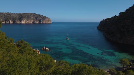 Sailing-boat-yacht,-clear-turquoise-blue-sea-water,-trees,-remote-natural-bay,-Palma-de-Mallorca-Island