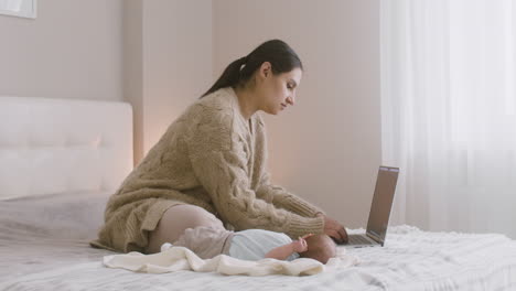 Junge-Mutter-Sitzt-Auf-Dem-Bett-Und-Arbeitet-Am-Laptop,-Während-Ihr-Neugeborenes-Baby-Neben-Ihr-Liegt