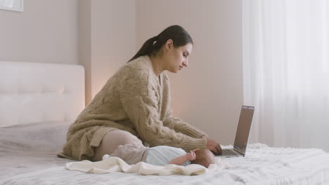 young mother sitting on the bed and working on laptop computer while her newborn baby lying next to her