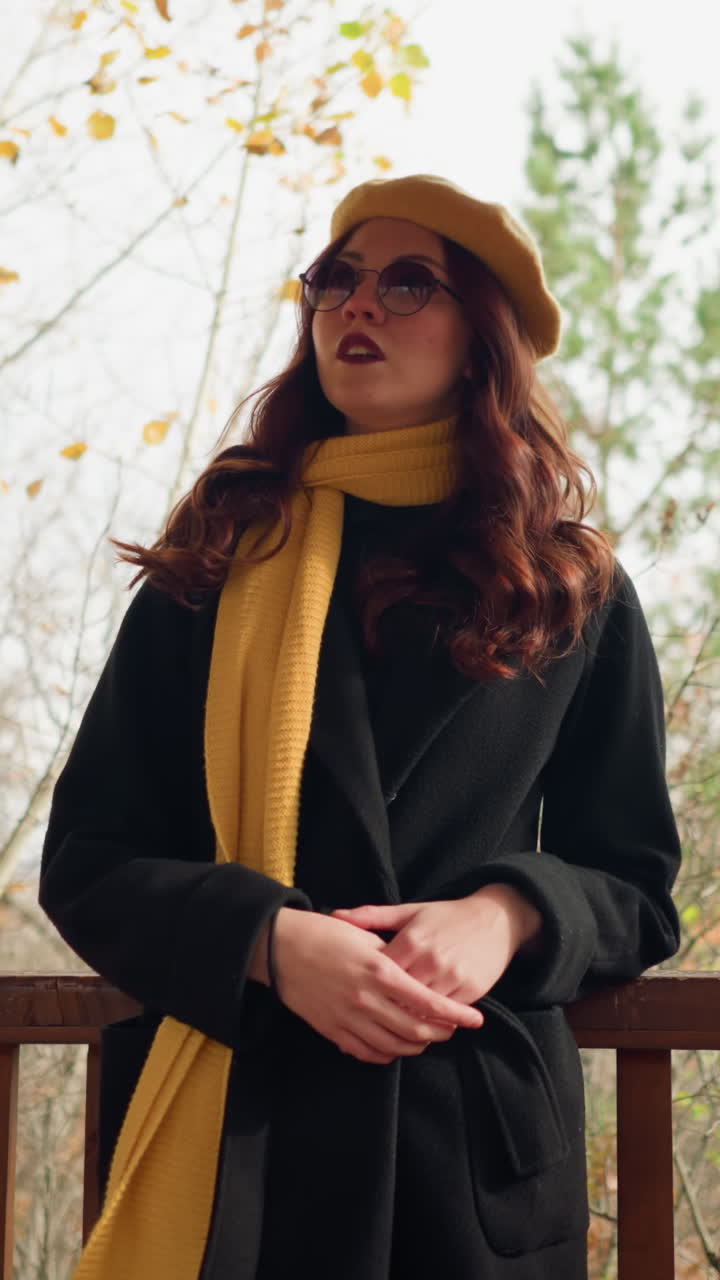 elegante dama en boina amarilla y silenciador descansa en la barandilla del mirador con la mano cerrada juntos, ajustando el cabello mientras mira a su alrededor en un entorno boscoso de otoño