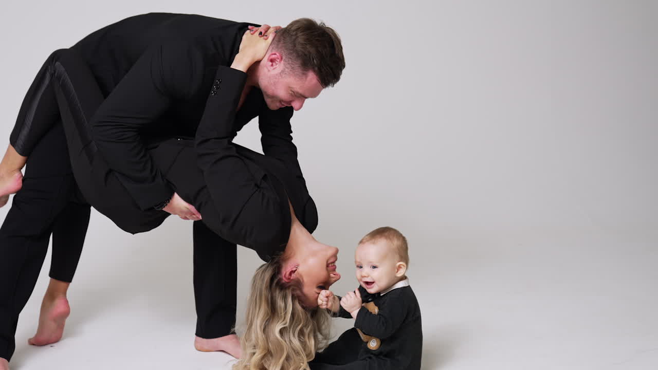 Man holding a woman bent behind. Little baby sitting on the floor touches the long blond hair of his mom. Studio footage of a family of three.