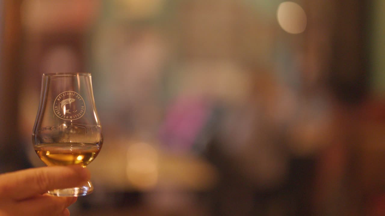 A hand gently swirls a whisky glass in a warmly lit pub or bar, with a softly blurred background and inviting, intimate atmosphere