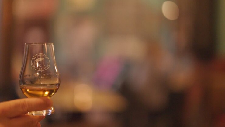 Una mano agita delicatamente un bicchiere di whisky in un pub o bar illuminato caldamente, con uno sfondo leggermente sfocato e un'atmosfera invitante e intima.